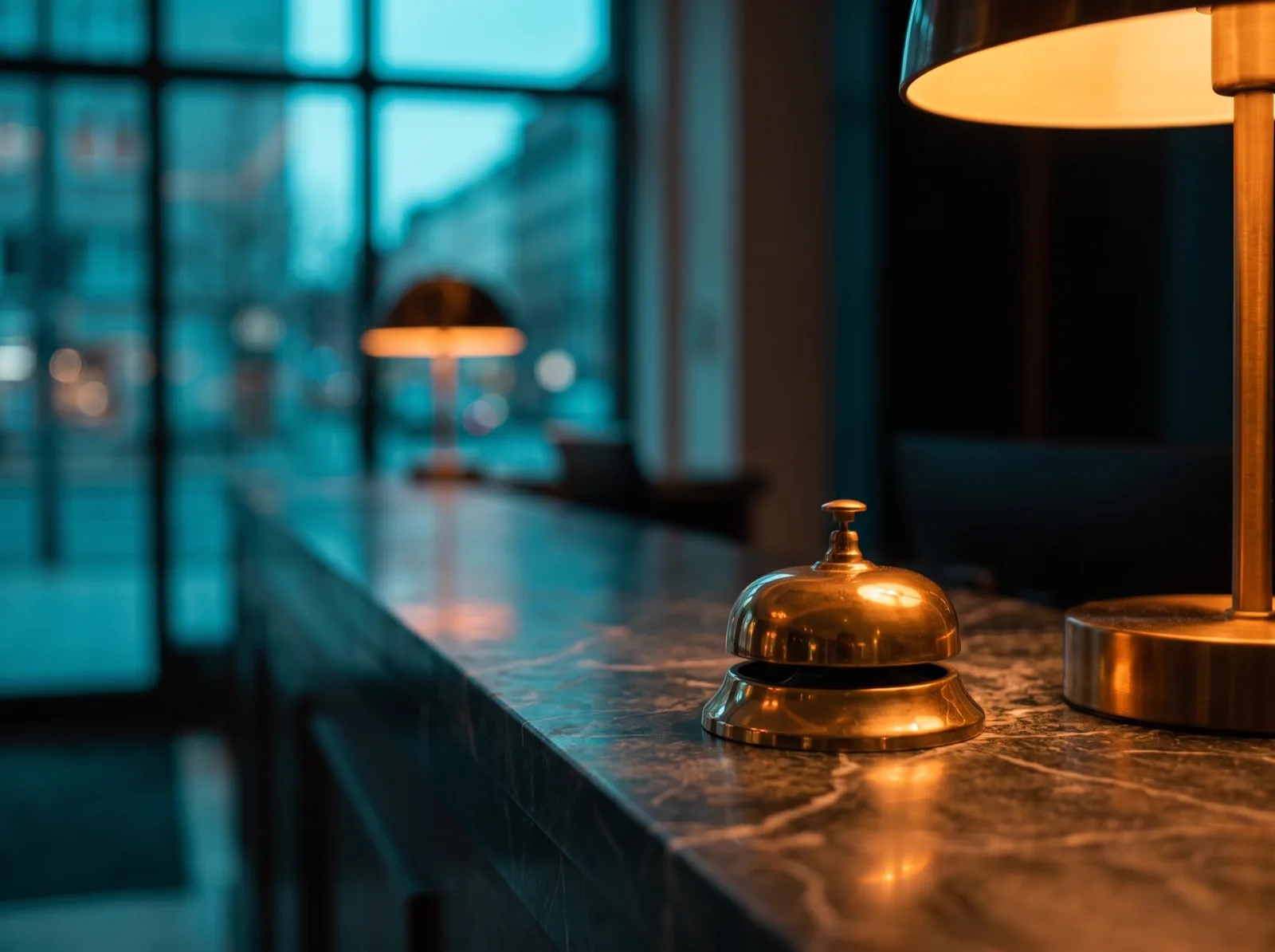 A polished brass service bell on a dark marble hotel counter, warm lamplight glowing around it, cool teal light from a lobby window behind